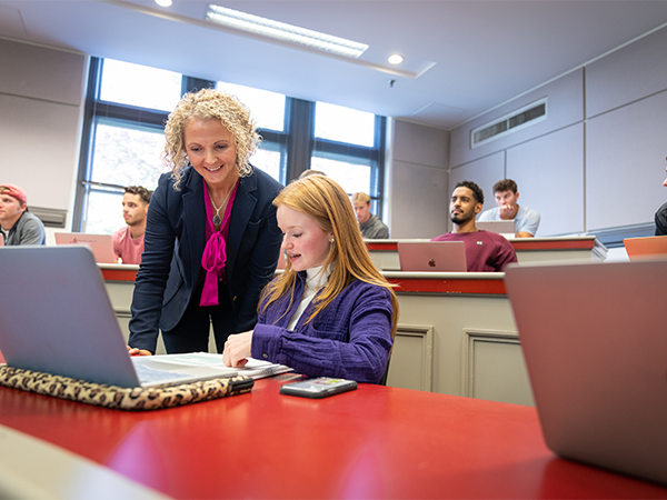 A professor leans over the shoulder of a student looking at a packet of materials in an accounting classroom.